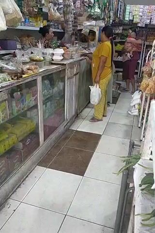 The video shows the interior of a small, cluttered convenience store. In the foreground, a woman in a yellow shirt and patterned pants stands at a counter, holding a white plastic bag. She is facing a woman behind the counter who is wearing a dark shirt and looking at her. The counter is filled with various items, including packaged goods and what appear to be food containers. To the right of the first woman, another woman is visible, holding a baby. She is wearing a dark maroon shirt and dark pants. Behind the counter, shelves are stocked with more merchandise, including bags of snacks and various packaged goods. The floor is tiled with white and brown squares. The lighting appears to be artificial and somewhat dim, typical of an indoor retail environment. The overall impression is of a busy, everyday transaction taking place in a local shop.