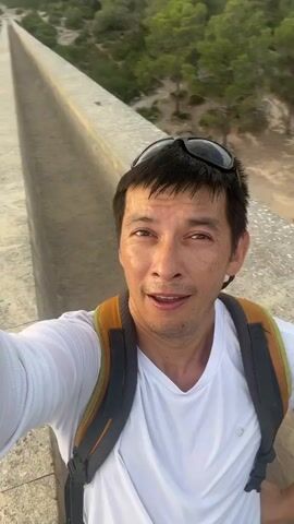 A man is filming himself with a smartphone. He is wearing a white t-shirt and a backpack. He has sunglasses on top of his head. The man is standing on a stone structure, possibly a bridge or aqueduct, with trees and a hill in the background. The camera angle suggests he is holding the phone out in front of him. The man is looking at the camera and appears to be speaking or reacting to something. The lighting suggests it is daytime, possibly late afternoon.