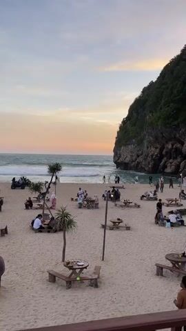 The video shows a wide shot of a crowded beach with picnic tables scattered on the sand. The ocean is visible in the background, with gentle waves rolling in. A large, green, tree-covered cliff dominates the right side of the frame. People are seated at the tables, some eating and others chatting. Several individuals are walking on the beach, and a few are gathered near the water's edge. The sky is a soft gradient of orange and blue, indicating either sunrise or sunset. The overall atmosphere appears relaxed and social.