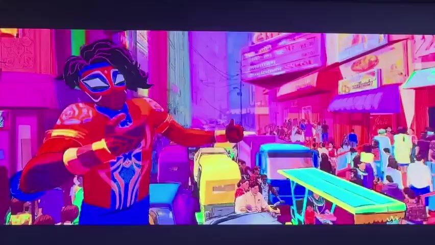 The media file displays a vibrant, animated scene set in a bustling city, likely in a location with a diverse architectural style, given the blend of modern and older structures. The primary subject is a character dressed in a red and blue costume with spider-like motifs, suggesting a superhero or a character inspired by such figures. This character appears to be in motion, possibly performing actions related to their abilities or interacting with the environment.

The setting is a city street filled with various vehicles, including cars and what appear to be auto-rickshaws or similar small, colorful vehicles. Pedestrians are also visible, adding to the sense of a lively urban environment. The city skyline features buildings with domes and intricate facades, alongside more contemporary structures. The overall color palette is striking, with intense blues, purples, pinks, and yellows dominating the scene, creating a surreal and dreamlike atmosphere. The lighting suggests it could be daytime, but the exaggerated colors give it an unnatural feel.

The character's actions, such as pointing and gesturing, imply a narrative or an ongoing event within the scene. The character's facial features, though stylized, convey a sense of focus or determination. The visual style is reminiscent of modern animated films, with bold outlines and a high level of detail in the character and background designs. The presence of multiple vehicles and people suggests a public space, possibly a busy market or main thoroughfare. The overall impression is of a dynamic and visually rich scene, possibly from a film or video game. No text is immediately discernible in the provided frames.