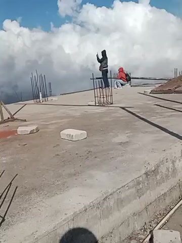 A construction site with a concrete slab and rebar is shown. Two people are present. One person is standing and appears to be taking a photo with a phone. The second person is sitting. The site is at a high elevation, with clouds visible below and mountains in the distance. The sky is blue with white clouds.