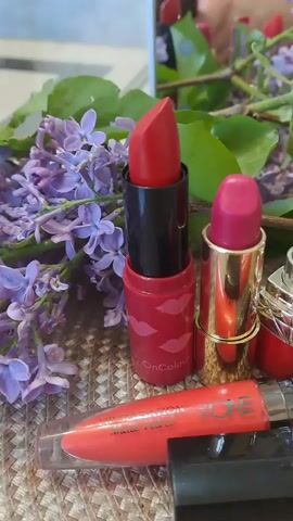 The video shows a collection of lipsticks and lip glosses displayed on a woven surface, surrounded by purple lilac flowers. The lipsticks are various shades of red and pink, with different packaging including gold, silver, and black casings. The lip glosses are in clear tubes with red and black caps, labeled with "mark." and "ONE". A blurred reflection of the person holding the camera is visible in the background, suggesting the shot is taken close-up. The overall lighting is soft and natural, enhancing the colors of the makeup and flowers.
