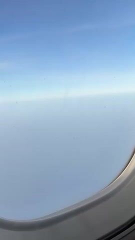 The video starts with a view from an airplane window, showing a mostly cloudy sky. The camera then pans to show the interior of the airplane cabin. Overhead bins are visible, along with the ceiling lights. The plane appears to be at least partially full with passengers seated.