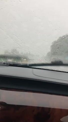 The video is shot from inside a car, looking out the windshield. It is raining heavily, with water droplets visible on the glass, obscuring the view of the road ahead. The road is wet and appears to have some standing water. Other cars are visible in the distance, with their headlights and taillights illuminated. The surrounding environment is green with trees and foliage, but the heavy rain and fog make it difficult to discern details. The interior of the car shows a portion of the dashboard, which has a dark color and a wood-grain trim. The overall impression is one of driving in a downpour.