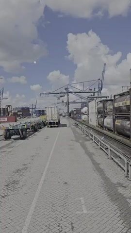 A long shot video shows a view of a shipping yard on a cloudy day. In the foreground, there is a paved area with some empty trailers on the left and a white shipping container on a truck in the middle of the road. To the right, there are train tracks with several tank containers on them. In the background, there are several large cranes and more shipping containers, suggesting an active port or distribution center. The sky is blue with scattered white clouds.
