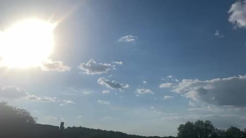 The video shows a bright, sunny sky with scattered white clouds. The sun is visible on the left side of the frame, shining intensely. In the distance, a treeline forms a dark silhouette against the sky. The lower portion of the frame reveals a dark structure, possibly a fence or wall, and some greenery. The overall impression is of an outdoor scene on a clear day.