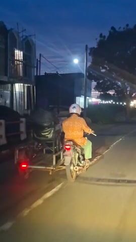 A person rides a dark-colored motorcycle equipped with a homemade, dark-colored sidecar or trailer carrying a large, dark, lumpy object. The rider wears an orange patterned shirt, light green pants, and a light-colored helmet, and is moving away from the camera. The scene is illuminated at night by streetlights and the presumed headlights of the camera's source. On the left side of the asphalt road, multi-story buildings are visible, some with illuminated windows, and a white and red barrier is present. The sky is dark blue.