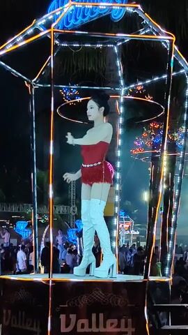A woman dances on an elevated platform at an outdoor event. She is wearing a red strapless dress and white thigh-high platform boots. The platform is a lighted, metal framed structure with the word "Valley" at the base. Many people are present in the background. Some appear to be watching the woman dance while others are gathered in groups. The event appears to be at night as the sky is dark. A palm tree can be seen in the background, as well as various decorative lights.