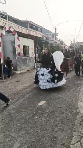 A person in a black and white ruffled costume with large white wings walks down a dirt road, holding a staff. The person is wearing a crown and has their hair pulled back. The costume has a layered, voluminous skirt. In the background, there are houses, a makeshift arch decorated with red and white flags, and other people watching. The weather appears to be overcast. The person is walking towards the right of the frame.