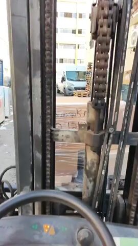 The video shows a first-person perspective of someone operating a forklift. The operator's hand is visible on the steering wheel. The forklift is in an outdoor industrial area, possibly a warehouse or loading dock. Pallets of goods wrapped in plastic are visible in the background, along with other vehicles and buildings. The forklift's control panel with buttons and a digital display is also visible. The video appears to capture the initial moments of operation.