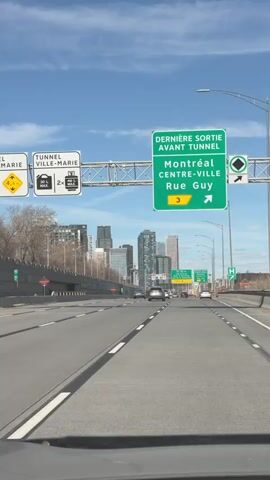 The video shows a first-person perspective from inside a vehicle driving on a highway towards a cityscape. The road is wide with multiple lanes, marked with white lines and black dividers. Several cars are visible on the road, traveling in the same direction. Overhead, green highway signs indicate directions to Montreal's downtown and Rue Guy. Additional signs warn about the upcoming tunnel. The cityscape is visible in the distance under a blue sky with scattered clouds. Street lights line the side of the highway.