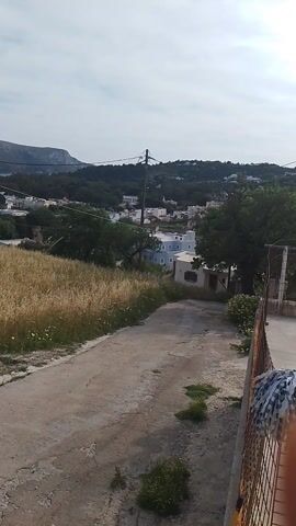 The video shows a view of a coastal town from an elevated position. A narrow, cracked pavement path runs along the right edge of the frame, with patches of greenery growing through the cracks. Next to the path, there's a field of dry, golden grass, indicating a warm climate. In the distance, a small town with white buildings is nestled between a forested hillside and the coastline. The sea is visible in the background, and a large mountain rises on the left side of the frame. Power lines stretch across the scene, slightly obscuring the view of the town. The sky is overcast, with a hazy appearance. The camera remains still throughout the video.