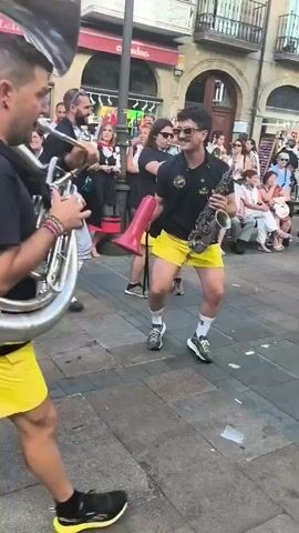 The video shows a street performance with several musicians. In the foreground, a person wearing a black t-shirt and bright yellow shorts is playing a sousaphone. Behind them, another person in similar attire plays a saxophone. A third individual in the background is playing a trumpet and holding a megaphone. The musicians are surrounded by a crowd of people, some of whom are wearing party hats. The environment appears to be a European city street with buildings lining both sides and cobblestone pavement. There is confetti scattered on the ground. The overall atmosphere is festive and celebratory.