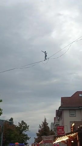 A person walks on a tightrope high above the ground, with buildings and trees visible below. The sky is cloudy. The person is wearing a white shirt and blue pants and is holding their arms out for balance. The tightrope is suspended between two points, and the person is moving from left to right in the frame. There are also some stalls or tents visible below, suggesting a fair or market. The video is shot from a low angle, looking up at the tightrope walker.