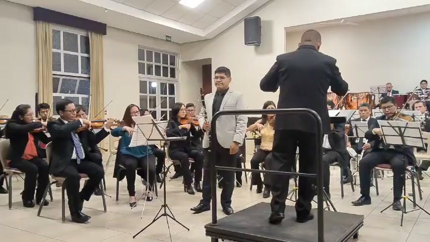 An orchestra is performing in a large, brightly lit room. The orchestra consists of violins, cellos, a clarinet, a drum set, and other instruments. The musicians are dressed in formal attire. The conductor stands on a small platform and directs the orchestra with sweeping arm movements. The clarinet player stands to the conductor's left, also on the platform. The walls are light-colored, and there are large windows on one side of the room. There is a speaker mounted high on the wall. There are music stands in front of each musician.