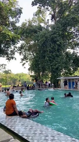 The video shows a lively outdoor swimming pool scene on a sunny day, surrounded by lush greenery and tall trees. A diverse group of people, including children and adults, are enjoying the water. Some are swimming, while others are relaxing on inflatable rings or floating devices. In the foreground, a person wearing an orange shirt is seated on the edge of the pool, facing away from the camera, with another person lying down next to them. In the background, more people are gathered around the pool, some standing and conversing, others observing. A small building, possibly changing rooms or restrooms, is visible on the right side of the frame, with people entering and exiting. The water in the pool is clear and turquoise, reflecting the bright sky. The overall atmosphere is one of recreation and relaxation.