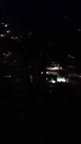 THE VIDEO IS TAKEN AT NIGHT SHOWING A VERY BLURRY AND DARK SCENE. THE BACKGROUND APPEARS TO BE A CITY OR TOWN WITH NUMEROUS LIGHTS SCATTERED THROUGHOUT. IN THE FOREGROUND, THERE IS A VERY BLURRY, DARK AREA WITH A FEW LIGHTS VISIBLE. THE OVERALL QUALITY OF THE VIDEO IS VERY POOR, MAKING IT DIFFICULT TO DISCERN ANY SPECIFIC DETAILS ABOUT THE LOCATION OR ANY OBJECTS OR PEOPLE PRESENT.