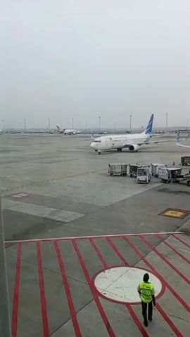 A wide-angle shot of an airport tarmac under an overcast sky. In the foreground, a person in a bright yellow-green vest walks away from the camera on a red-lined platform. In the mid-ground, a white airplane with blue and green accents on its tail is parked on the tarmac. The plane has "GARUDA INDONESIA" written on its side. Several baggage carts and service vehicles are positioned near the airplane. In the background, other airplanes are visible on the tarmac, and a long row of airport buildings stretches across the horizon. The overall impression is one of a busy, operational airport.