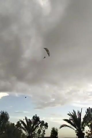 The video shows two paragliders soaring through the sky. The sky is filled with puffy, gray clouds, although there are patches of blue visible. In the foreground, trees and buildings are silhouetted against the sky. The paragliders are seen at a distance; one is higher than the other. The higher one is easier to see in greater detail.  The camera zooms in on the higher paraglider to provide a better view.