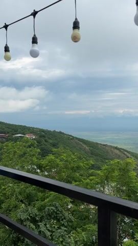 The video is an exterior shot taken from a balcony or elevated vantage point, looking out over a lush green landscape. Hanging lights are visible in the foreground, suspended from a wire. The landscape consists of rolling hills covered in dense green trees, with a few buildings visible on the slopes. In the distance, a wide valley or plain stretches out, with mountains or a range of hills visible on the horizon. The sky is cloudy, with a mix of light and dark clouds, suggesting an overcast or potentially rainy day. The overall impression is of a scenic view from a high location. There are no people visible in the video.