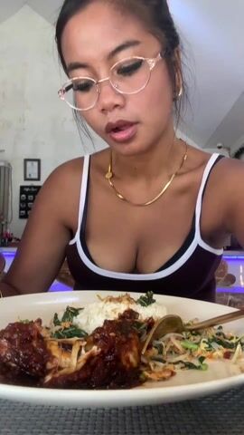 A woman with dark hair in a bun and glasses is eating food from a large plate in front of her. She is wearing a dark tank top with white trim and a gold chain. She takes a spoonful of greens from the plate and eats it. The plate contains rice, chicken with a dark red sauce, and a salad of sprouts. The setting is in a restaurant or cafe. There is a framed picture, a coffee menu, and a bar with a blue light in the background.
