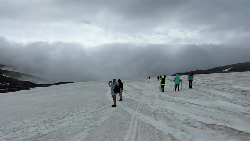 A group of people are walking on a snowy landscape under a cloudy sky. Some are taking pictures, while others are walking with hiking poles. The terrain is covered in snow with some patches of dirt and rock visible. In the distance, a thick layer of clouds obscures the mountains. The overall atmosphere is overcast and cool.