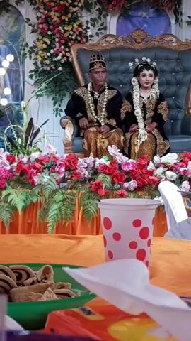 This is a video taken at an event, likely a wedding. In the background, a man and a woman are seated on a decorated throne, dressed in traditional attire. The setting is adorned with flowers, curtains, and lights, suggesting a festive occasion. In the foreground, there are snacks and drinks, with a focus on a jar filled with crackers and another container with a beverage. The camera pans and zooms, capturing different elements of the scene, including a glimpse of a stage with musical instruments and performers in the distance.