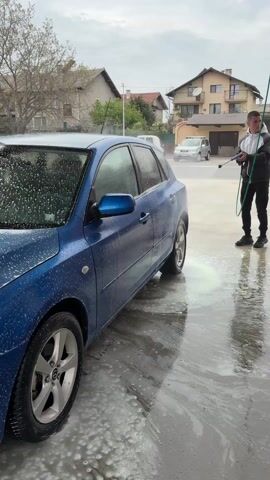Here is a concise description of the video:

The video shows a blue Mazda being power-washed at a car wash. A person wearing black pants and a grey hoodie is holding the power washer and spraying the car. The car is covered in soap, and the ground is wet. There are buildings visible in the background under a cloudy sky.