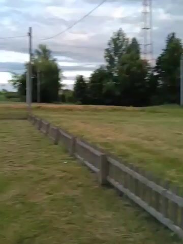 This video pans across an outdoor playground area in Smolino, Russia. The playground features a brightly colored climbing structure with a slide, a sandbox, and a swing set. Adjacent to the playground is an outdoor fitness area with several exercise machines. The scene is set in a grassy, open field with trees and buildings visible in the background. A wooden fence runs along the edge of the playground. The sky is cloudy, suggesting it might be late afternoon or early evening, and the lighting is dim. There are no people visible in the video. The overall mood is peaceful and serene, hinting at a quiet, possibly rural, community setting.