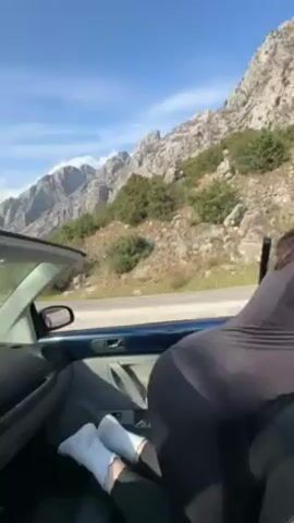 The video begins inside a parked, blue Volkswagen Beetle convertible, showing a woman mostly out of frame and facing the passenger side. The car is parked with a view of mountains in the background under a partially cloudy sky. The camera angle shifts to show the landscape from the perspective of the driver's seat. The camera then shows the car parked in front of a scenic view overlooking a body of water, with mountains visible in the distance and a bench nearby. The sun is shining through a bare tree in the foreground.