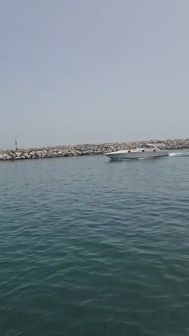 A white speedboat travels at speed across calm, dark blue water. The boat is oriented from left to right across the frame. A breakwater, constructed of large, rough grey stones, stretches horizontally across the background. A red and white striped marker pole is visible on the left side of the breakwater. The sky is a pale, hazy blue with no visible clouds. The boat is leaving a white wake behind it.