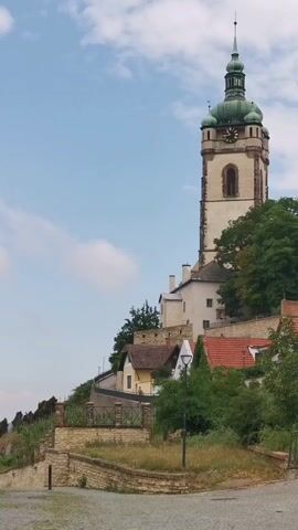 A video clip shows a historical tower on a hillside with a cloudy sky. There are several buildings with red roofs clustered on the slope below the tower, partially obscured by trees and greenery. A section of a stone wall and stairs is visible in the foreground. A person's head and shoulders, wearing a patterned shirt, are visible in the lower left corner of the frame, facing away from the camera towards the scene. The tower has a white facade, a green steeple, and a clock face.