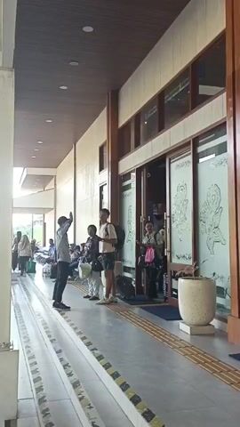 A group of people are standing and sitting in a covered outdoor walkway or waiting area. The walkway has a wooden ceiling and a tiled floor with a yellow and black striped border. On the right side of the walkway, there is a glass-doored building with decorative etchings on the glass. People are standing near the entrance of the building, some with luggage. One person in a grey uniform is talking to a person in a black shirt and shorts who is holding a backpack. Another person in a green uniform is standing to the right of the person in the black shirt. In the background, there are more people sitting on benches. The lighting suggests it is daytime.