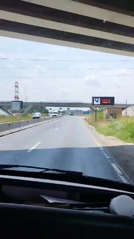 The video shows a first-person perspective of someone driving on a highway. The road is paved with white lane markings. To the left of the highway, there is a concrete barrier and a white van driving in the same direction. Further in the distance, there are several other vehicles and what appears to be a train track elevated above the highway. A pedestrian bridge crosses over the highway. On the right side of the highway, there is a billboard with an advertisement and some vegetation. The sky is partly cloudy. The dashboard of the vehicle is visible in the foreground, with some papers and a white object on it.