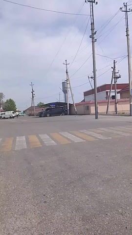 The video shows a large, white mosque with a dome and minarets in a city. The mosque is located on a street corner near a building that houses a market. Cars drive by and people can be seen walking on the sidewalk near the mosque. The video appears to be taken from a moving vehicle.