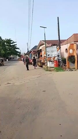 The video captures a street scene in Ohokobe Ndume, Nigeria, under a bright, clear sky. The paved road, showing visible cracks and some debris, leads into the distance. Two individuals dressed in dark clothing are walking away from the camera on the left side of the street. Further down, a red, open-backed vehicle or cart is visible, along with other pedestrians.

On the right side of the road, a row of single-story buildings, likely shops or small businesses, lines the street. These structures exhibit varying colors like orange and brown, with some featuring stalls or displays outside. An open drainage ditch runs along the front of these buildings. A modern street light with a solar panel stands prominently on the right, and multiple utility poles with overhead wires crisscross the upper part of the frame. A large sign with indistinct text and images is attached to one of the buildings, and several red barrels and white containers are placed nearby. A large tree with green foliage is visible on the far left. The scene depicts typical daytime activity in a bustling street environment.