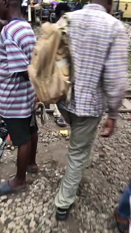 The video shows a busy outdoor market with several people walking on and around train tracks. The ground is covered in gravel and dirt. In the foreground, a person wearing a pink tank top and khaki pants walks away from the camera, carrying a large beige backpack. To the left, a person in a striped shirt walks away from the camera. To the right, a person in a plaid shirt walks towards the camera. Further in the background, various stalls are set up, with people browsing and selling goods. There are umbrellas providing shade over some of the stalls. The overall impression is a vibrant and active marketplace situated on active train tracks.