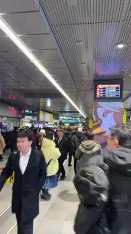 The video shows a crowded subway station hallway with people walking in both directions. The hallway has a grey corrugated metal ceiling with long, thin fluorescent lights. There is a large digital display showing train schedules and advertising. People of various ages and dressed in a mix of business attire and casual wear are walking. Many are carrying bags or briefcases. The floor is light-colored tile with yellow tactile paving.