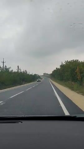 The video shows a view from inside a car looking out at a two-lane asphalt road. The sky is overcast and gray, suggesting it might be raining or has recently rained, as there are water droplets visible on the windshield. The road curves to the right in the distance. On the left side of the road, there are some power lines and trees. On the right side of the road, there is a sandy shoulder and dense green trees. A dark-colored car is visible in the distance, driving in the same direction as the camera car. The car appears to be on the left lane, which is for oncoming traffic in the US but could be for the same direction of travel in other countries. The video captures the movement of the car along the road.