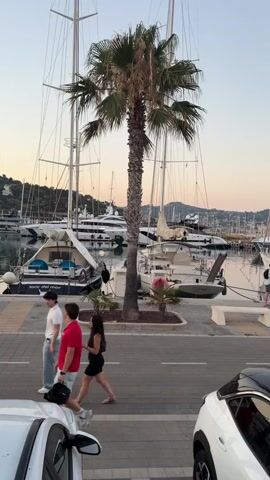 This media file captures a serene evening scene at a marina, likely in Imperia, Italy. The primary subjects are several sailboats and yachts docked in the harbor, their masts reaching towards a pale, dusky sky. A prominent palm tree stands tall in the foreground, its fronds rustling gently.

Several people are visible walking along the paved waterfront promenade. A group of three individuals, two men and a woman, are walking from left to right. Further down the promenade, a woman and a child are seen walking. The presence of these individuals suggests a leisurely stroll or an evening outing.

The time of day appears to be dusk or early evening, indicated by the soft, warm light and the pastel colors in the sky. The weather seems pleasant, with no signs of rain or strong winds. The setting is clearly a marina, with numerous boats of varying sizes moored. In the background, hills and buildings can be seen, providing a picturesque backdrop.

Notable details include a white bench placed near the palm tree, offering a spot for rest. The text "FORO DEL MAR" is visible on the side of a boat, which could be the name of the boat or a location indicator. The overall mood conveyed is peaceful and relaxed, typical of a Mediterranean evening by the sea. Two cars are parked in the foreground, one white and another partially visible.