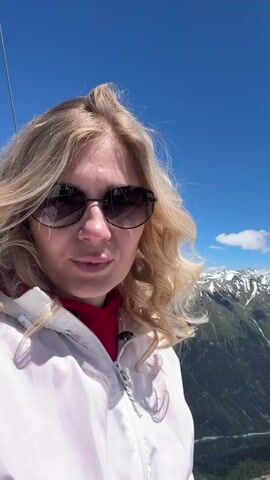 A blonde woman wearing sunglasses and a white jacket is taking a selfie on a mountain. She is smiling at the camera, and behind her, snow-capped mountains rise under a clear blue sky. The woman is standing on a rocky ledge, with some snow patches visible. The video pans slightly to show the vast mountain range and a glimpse of a structure with red accents and cables, possibly part of a ski lift or observation deck.