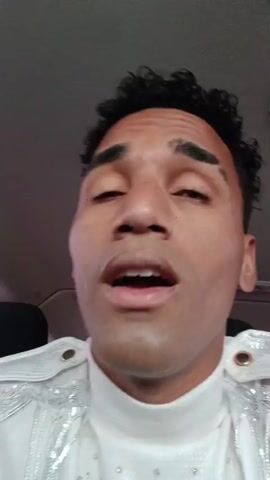A man with dark curly hair and a white sequined jacket records himself with a phone camera while sitting in a vehicle. He talks to the camera, and in the background, a person with glasses and a black and white striped shirt flashes a peace sign. The vehicle appears to be moving.