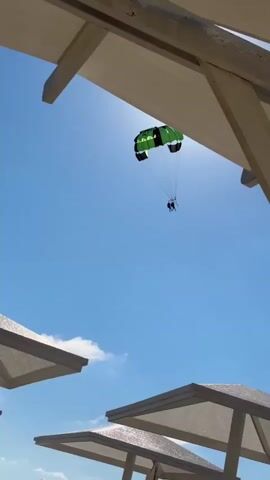 A parasailer with a green and black parachute is visible against a clear blue sky. Two people are suspended beneath the parachute. The parasailer is moving from left to right in the frame. In the foreground, there are white beach umbrellas and lounge chairs. The sun is shining brightly, casting shadows and creating a glare. The scene appears to be at a beach or coastal resort.