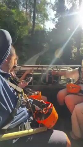 A group of people are riding in the back of a truck, seemingly headed towards a ziplining adventure. They are wearing harnesses and have orange helmets. The truck is driving on a dirt road through a forest with tall trees and the sun shining brightly. Some people are holding phones.