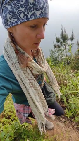 A woman in hiking attire stops on a hillside trail. She wears a blue and white patterned head wrap, a green long-sleeved shirt, a patterned scarf, and hiking shoes. A plaid shirt is tied around her waist. Sunglasses are perched on her head. The woman kneels down to examine a patch of small white flowers. The hillside is covered with greenery, including trees and tall grasses. Mist hangs in the air.