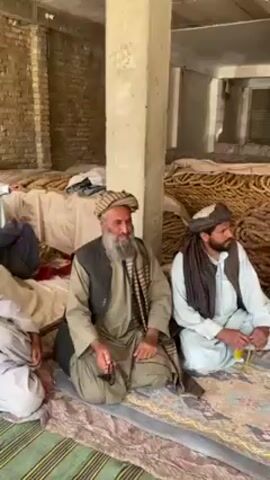 Several men sit cross-legged on a rug in an indoor area. They wear traditional clothing, including head coverings, and vests. Some men have beards. They are talking and laughing with each other. In the background are large piles of coiled material. The room has brick walls and concrete pillars.