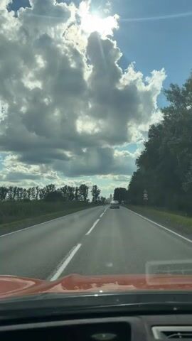 A video taken from inside a vehicle driving on a paved road. The road has two lanes and a solid white line dividing them. The sky is blue with large, fluffy white and gray clouds. The sun is shining brightly through a gap in the clouds, creating a halo effect. On the left side of the road, there is a field of trees. On the right side of the road, there are more trees and a road sign. There are a few other vehicles visible on the road ahead. The front of the vehicle in which the video is being filmed is visible in the foreground.