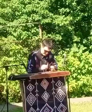 A person is playing a dulcimer outdoors. The person is wearing sunglasses and a black jacket with white and colorful accents. The dulcimer is on a stand covered with a black cloth that has a diamond pattern in white, red, and grey. There are green trees and a fence in the background. A microphone stand is visible to the left. The person is holding mallets in each hand and striking the strings of the dulcimer. The video is comprised of four frames, each about one second long, showing the person playing the instrument.