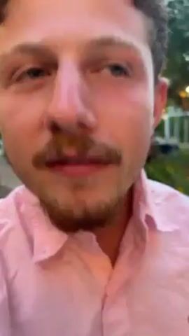 A close-up shot shows a person with curly brown hair, a mustache, and a short beard, wearing a light pink collared shirt. The person looks directly at the camera, with the background showing blurry greenery, what looks like a rock wall, and a few light fixtures. The person initially moves their hand in front of the camera, then pulls it away to reveal their face.