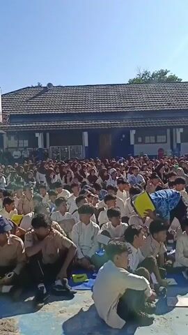 Here is a description of the video:

The video shows a large group of students sitting on the ground in an outdoor schoolyard or courtyard. The students are mostly boys dressed in school uniforms, and they appear to be watching a presentation or ceremony taking place in front of them. 

In the foreground, there are some students sitting with their backs to the camera, while the rest of the group faces toward the front. In the background, there is a building with a tiled roof and windows. A basketball hoop is visible. The sky is clear and blue, suggesting it is a sunny day.

Later in the video, two individuals, a man with a microphone and a woman wearing a hijab, stand in front of the crowd. Another man stands nearby. They are positioned behind a table covered with wrapped gift-like objects. The setting appears to be a school event or gathering.